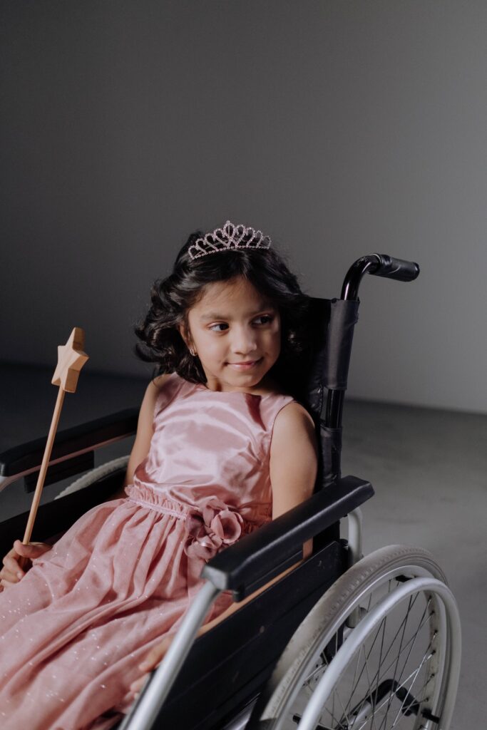 A little girl in a pink wheelchair with a tiara.